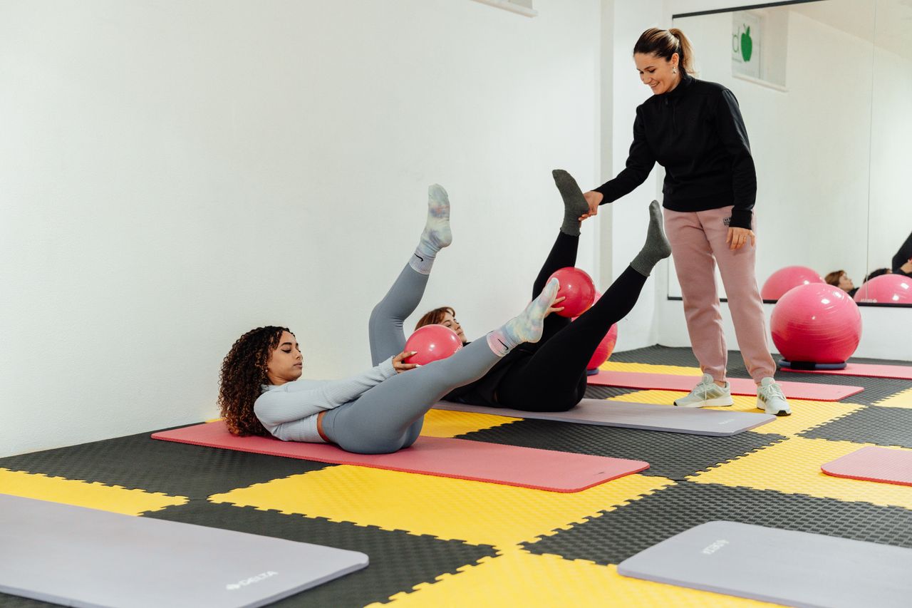 social-feed-img-02 Women engaged in pilates class using pink balls and yoga mats indoors at a gym.
