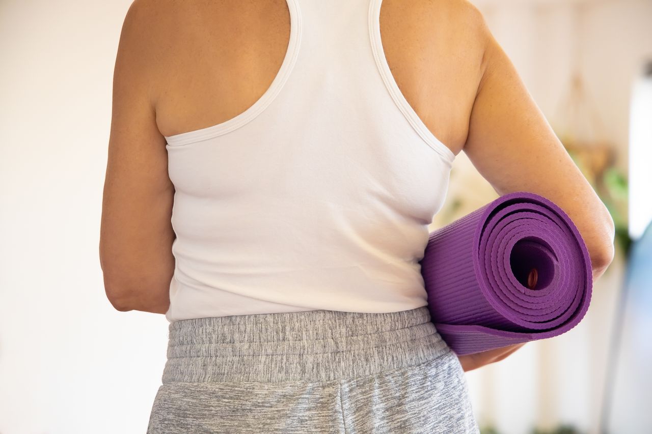 about-img Woman in white tank top holding a rolled purple yoga mat indoors, preparing for exercise.
