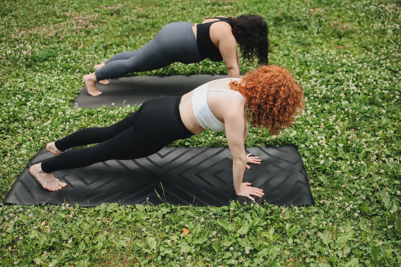 quality-assurance-02 Two women holding a plank position on yoga mats outdoors in a lush green setting.