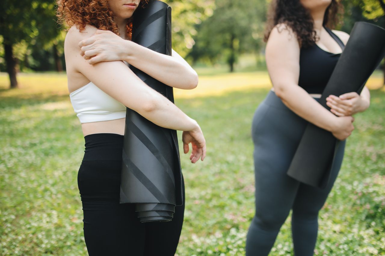 social-feed-img-05 Two women stand in a park holding yoga mats, preparing for outdoor exercise.